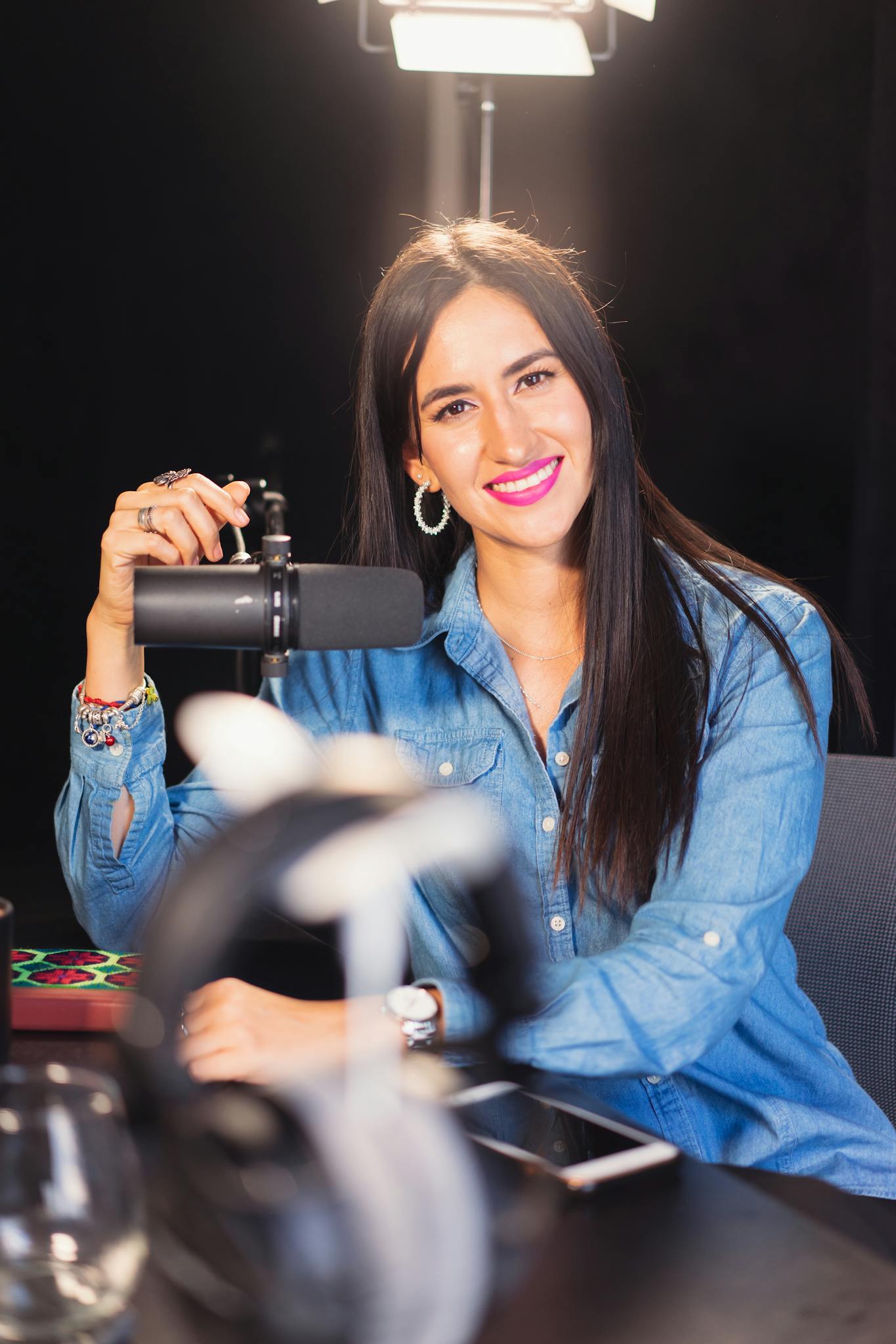 A cheerful woman wearing a denim shirt sits in a recording studio with a microphone.