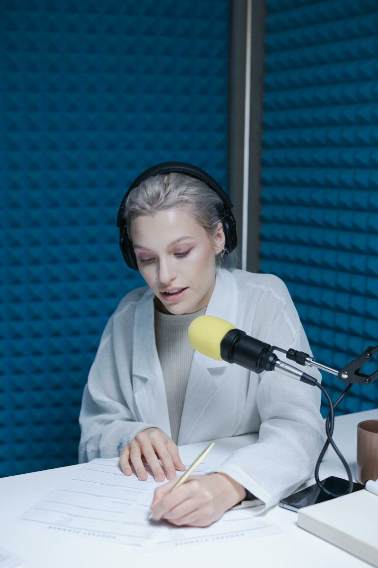 Woman in soundproof studio writing notes, wearing headphones, with microphone setup.